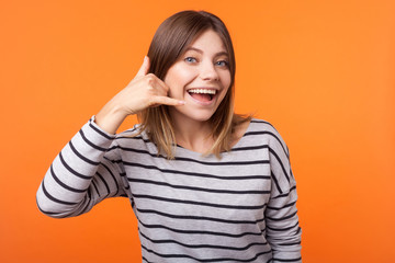 Call me! Portrait of cheerful beautiful woman with brown hair in long sleeve striped shirt standing, making call gesture with hand, looking playful. indoor studio shot isolated on orange background