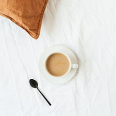 Cup of coffee with milk, black spoon, ginger pillow on white linen in bed. Minimalist morning breakfast concept. Flat lay, top view.