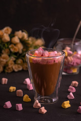 Glass cup with a double bottom with coffee with pastel colored marshmallows in the shape of hearts standing on a dark table with flowers.