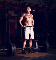 Obraz na płótnie Canvas Handsome concentrated brutal man standing at the barbell in crossfit gym on dark sport club background with shadows. Low section