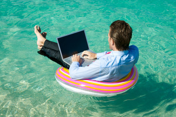 Relaxed businessman floating on a rubber ring while typing on his laptop