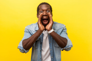 Attention! Portrait of scared man in denim shirt holding hands near wide open mouth and shouting...
