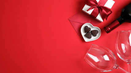 Top view table setting for Valentines day with bottle of wine champagne, two glasses, gift box with red ribbon bow, heart shaped candies and envelope on red background. Romantic dinner couple concept