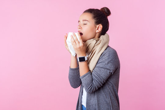 Influenza. Portrait of unhealthy brunette teenage girl with bun hairstyle wearing pullover and scarf sneezing into napkin feeling unwell, flu symptoms. indoor studio shot isolated on pink background