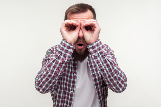 Portrait of curious wondered bearded man in casual plaid shirt looking through fingers gesturing binoculars, amazed surprised by what he discovers. indoor studio shot isolated on white background