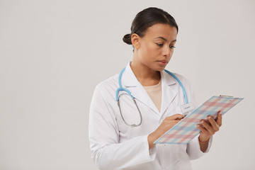 Waist up portrait of young mixed race nurse or female doctor holding clipboard while standing against white background, copy space