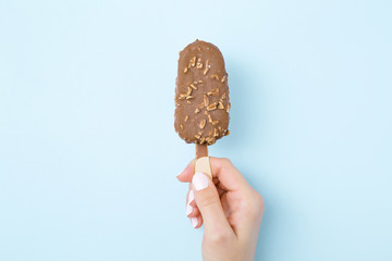 Young woman hand holding ice cream with nuts and chocolate glaze on light pastel blue table background. Point of view shot. Closeup.