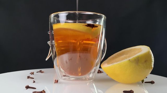 Adding sweet honey into a warm tea with lemon juice and cloves. Studio shot of isolated bees honey mixing with tea. A jar full of honey and half of lemon beside a cup of tea.