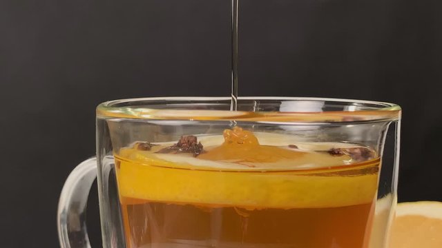 Adding sweet honey into a glass cup full of warm tea with lemon slice and cloves. Studio shot of isolated bees honey mixing with tea. Dark backdrop.
