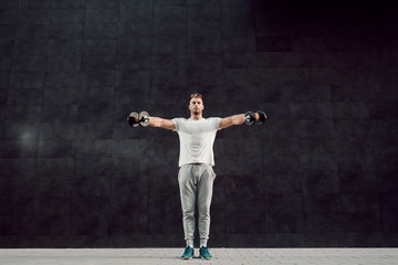 Fototapeta na wymiar Full length of attractive caucasian muscular bearded blonde man in tracksuit and with t-shirt standing in front of gray wall and holding dumbbells.
