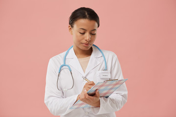 Waist up portrait of beautiful female doctor writing on clipboard while standing against pale pink background, copy space