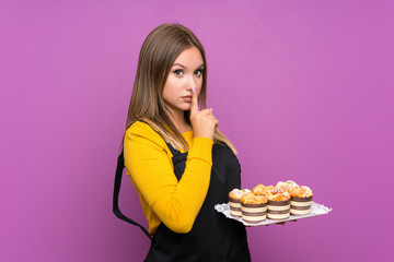 Teenager girl holding lots of different mini cakes over isolated purple background doing silence...