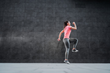 Side view of fit attractive Caucasian woman in sportswear and with ponytail running outdoors. In background is dark wall.
