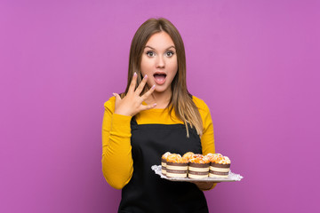 Teenager girl holding lots of different mini cakes over isolated purple background with surprise facial expression