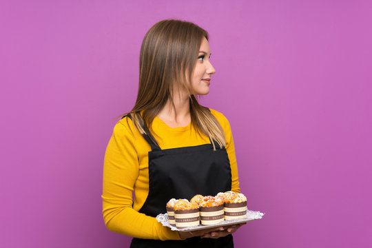 Teenager girl holding lots of different mini cakes over isolated purple background looking to the side