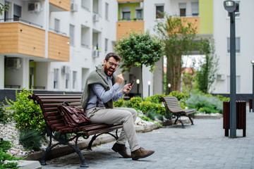 Successful handsome fashionable caucasian businessman sitting on bench in park, holding smart phone and rejoicing for achievement. Next to him is leather handbag.