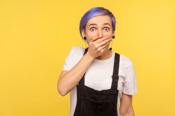 Portrait of scared terrified hipster girl with violet short hair in denim overalls covering mouth with hand and looking intimidated with eyes full of fear. isolated on yellow background, studio shot