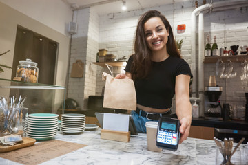 smiling barista woman standing behind the bar with hot coffee drink in paper cup and lunch paper bag for take away holding payment terminal