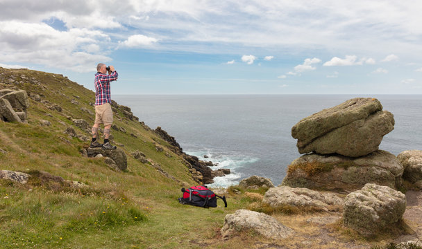 A hiker with binoculars looks out to sea at the Lands End lighthouse in south-west England. The man is standing on a rock on the high cliffs.