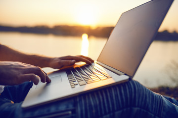 Close up of Caucasian man sitting in nature and using laptop. In background is river and sunset....