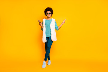 Full size photo positive afro american girl use smartphone browse promo point finger copyspace wear blue pants trousers teal jumper white vest stylish trendy isolated yellow color background