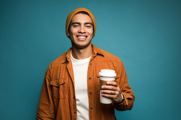 Recycling matters. Portrait of stylish mixed race man in yellow outfit, wearing hat, holding reusable plastic coffee or tea cup in hand and smiling isolated over blue background