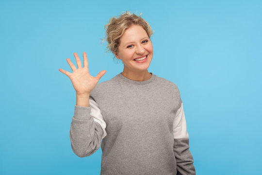 Hello! Cheerful woman with short hair in casual sweatshirt smiling friendly at camera and waving hi, welcoming or showing goodbye, hospitable greeting. indoor studio shot isolated on blue background