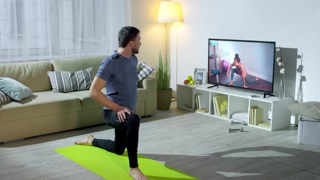 Medium shot of bearded man looking at female online personal trainer on TV screen and doing forward lunge to stretch legs