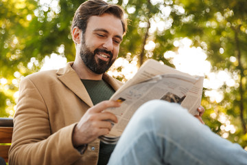 Image of handsome adult man reading newspaper on bench in park