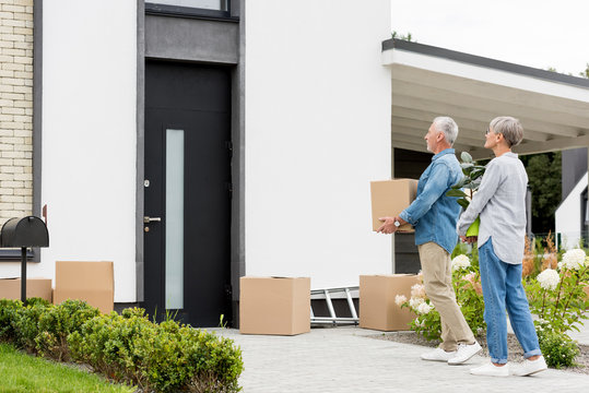 Mature Man Holding Box And Woman Holding Plant Near New House