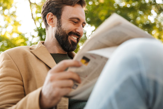 Image of handsome adult man reading newspaper on bench in park