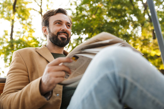 Image of handsome adult man reading newspaper on bench in park