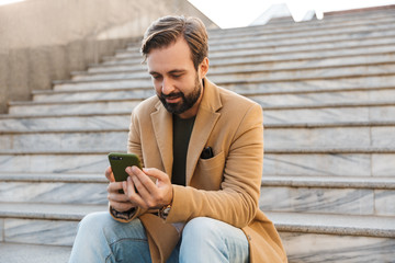 Image of handsome man holding smartphone while sitting on stairs outdoors