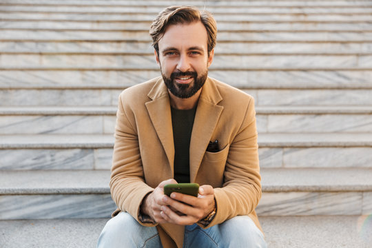 Image of handsome man holding smartphone while sitting on stairs outdoors