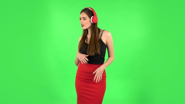 Cheerful girl dancing and enjoys music in big red headphones. Green screen
