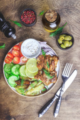 Lunch bowl with grilled chicken, red onion, tomato, cucumber, lemon, olive and sause on wooden background. Top view.