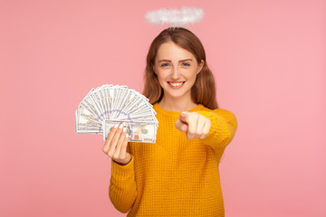 Hey you, make money! Portrait of positive angelic ginger girl with halo over head holding fan of dollar bills and pointing finger to camera, choosing lottery winner. indoor studio shot pink background