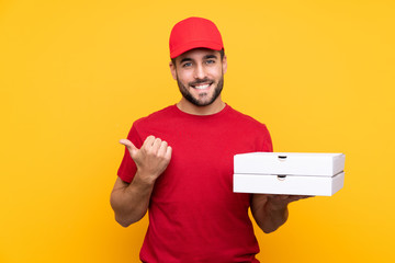 pizza delivery man with work uniform picking up pizza boxes over isolated yellow background pointing to the side to present a product