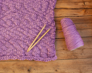 a knitted texture square purple fabric lies on a wooden table next to knitting needles and a skein...