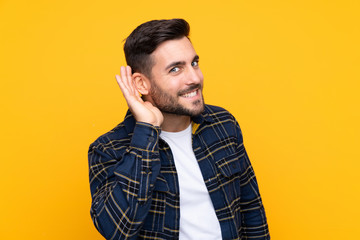 Young handsome man with beard over isolated yellow background listening to something by putting hand on the ear