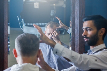 Perfect trim at barbershop. Young bearded man getting haircut by hairdresser with electric razor...