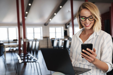Photo of happy blonde woman using cellphone and laptop while sitting