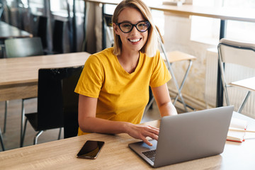 Photo of joyful young woman using laptop and smiling while sitting