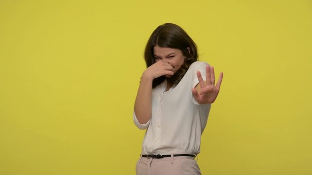 Bad smell! Displeased woman with brunette hair in blouse pinching her nose and showing stop gesture, feeling repulsion and disgust to stinky odor, fart. studio shot isolated on yellow background