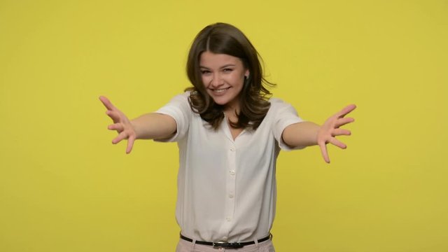 Come into my arms! Sincere kind hearted woman with brunette hair in blouse stretching hands to embrace you and looking at camera with friendly smile, free hugs. indoor studio shot, yellow background