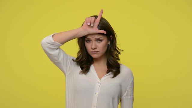 Frustrated woman with brunette hair in blouse standing with gloomy face and making loser gesture, snivelling crying, upset about failure, dismissal. indoor studio shot isolated on yellow background