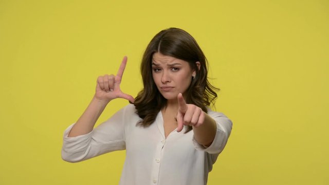 You are loser! Displeased woman with brunette hair in blouse showing loser gesture, L sign with fingers and pointing to camera, blaming for failure. indoor studio shot isolated on yellow background