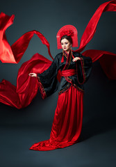 Woman is dressed in red Chinese Japanese folk clothing. Flying fabric, beautiful umbrella and fan in Japanese Chinese style, long earrings in the ears. Girl posing on a dark background