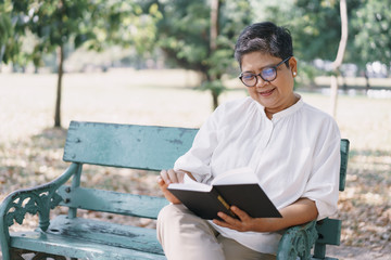 Happy elderly asian woman while sitting reading  book on the bench in the park. Concept of a happy lifestyle in the retirement