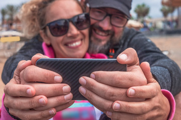 Two couple of hands of a middle-aged couple holding the mobile phone and smile for a selfie. Two attractive and relaxed people. Sitting on the beach on a winter vacation